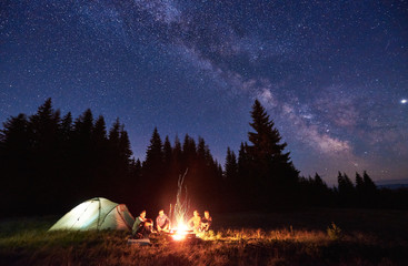 Night camping near bright fire in spruce forest under starry magical sky with milky way. Group of four friends sitting together around campfire, enjoying fresh air near tent. Tourism, camping concept. - obrazy, fototapety, plakaty