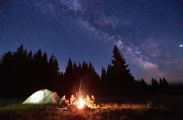 Cercles muraux Camping Camping de nuit près d& 39 un feu vif dans une forêt d& 39 épicéas sous un ciel magique étoilé avec voie lactée. Groupe de quatre amis assis ensemble autour d& 39 un feu de camp, profitant de l& 39 air frais près de la tente. Tourisme, concept de camping.