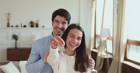 Proud happy young adult couple holding keys standing in own new apartment. Smiling husband and wife first time home owners real estate buyers looking at camera. Family mortgage investment concept
