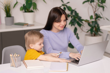 Cute son sitting at table and touching mothers laptop while assisting her with work, young mom working at home