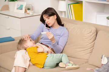 Fototapeta premium Worried young mother sitting on sofa and checking temperature of son while calling doctor by phone