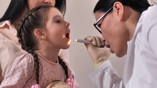 Close-up of cute preschool child opening mouth wide while positive asian female otolaryngologist examining her throat in medical clinic. Sick girl sitting on mom's knees during medical examination