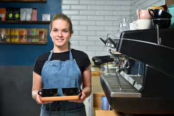 The bartender of the barista, a young girl in a coffee shop holds in her hands two mugs of coffee and smiles.