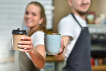 A young guy and a barista girl are standing with their backs to each other and holding a paper cup with coffee and a coffee mug. Small business and teamwork concept.