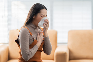  Asian woman sneezes. She uses a tissue to cover her mouth and she is working at home.