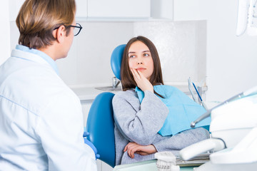 Young woman is sitting in dental blue chair in clinic, office. Girl is talking with man doctor in uniform before examination of teeth and treating. Visit to dentist, orthodontist concept.
