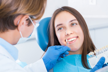 Young smiling woman is sitting in dental blue chair in clinic, office. Man doctor in gloves is selecting best color of implants using teeth samples. Visit to dentist, orthodontist concept.