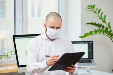 A man in a medical face mask against the coronavirus (COVID-19). An office worker making notes at his workspace with computers and green plants in the background. Coronavirus quarantine