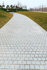 a neat concrete light tile path bends between fresh green grass in a new modern park on a cloudy day