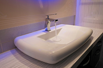 Close up shot of modern interior bathroom sink with blue mirror backlight. White ceramic washbasin in contemporary minimalism style. Copy space, background.