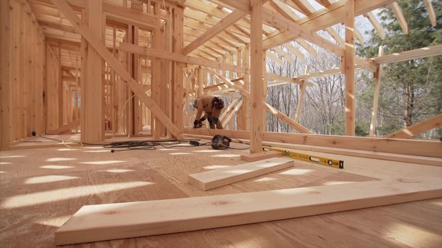 Builder in gloves is cutting wooden planks with small electric circular saw. Panoramic view of interior of spacious room in wooden frame house under construction