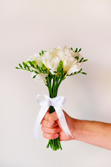 Cropped shot of man's hand holding beautiful bouquet of white freesia flowers tied with a white silk ribbon. Male handing the flowers as present. Close up, copy space, background.