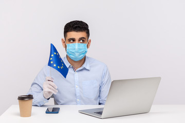 Pensive man in hygienic mask and gloves holding EU flag, looking worried and thoughtful, pondering to solve coronavirus problem, Covid-19 epidemic anxiety in Europe, quarantine. studio shot, indoor