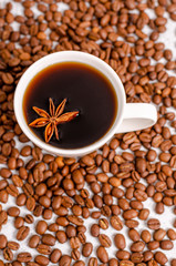 A cup of black aromatic coffee in a white cup with an anise star on a background of roasted arabica coffee beans on a light table.