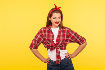 Portrait of positive pinup girl with bright makeup, red lipstick, in checkered shirt and headband, holding hands on hips