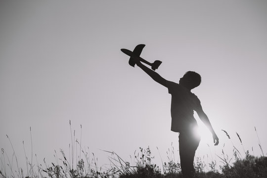Black silhouette of young caucasian kid isolated on sunny sunset sky background. Boy playing toy plane outside on grassy summer hill. Setting big goals and dreaming about happy future concept.