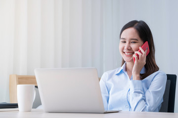 Attractive Asian woman works from home with laptop on desk in bedroom. Young girl smiling with happiness face while talking on phone. Telecomuting, freelancer and modern lifestyle concept.