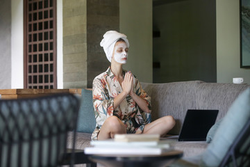 Young woman sitting on sofa with face beauty mask in front of laptop and doing yoga or meditation. Cozy time at home. Self time and home spa concept. Coronavirus outbreak and self-quarantining concept