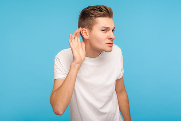 I can't hear. Portrait of curious nosy attentive man in white t-shirt holding hand near ear and listening carefully to gossip news, spying secrets. indoor studio shot isolated on blue background