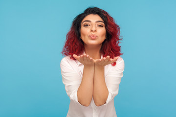 Portrait of happy fancy hipster woman in love holding palms up and sending sensual air kiss with pout lips, flirting to camera, expressing romantic feelings. studio shot isolated on blue background