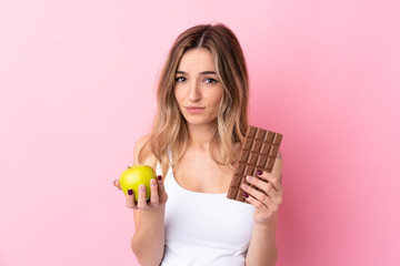 Young woman over isolated pink background taking a chocolate tablet in one hand and an apple in the other