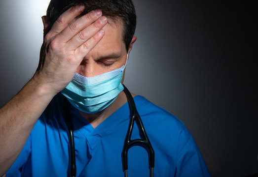 Stressed / tired doctor wearing blue hospital scrubs with surgical face mask, against a dark background.