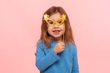 Portrait of fascinating cute little girl looking at camera through paper glasses and smiling, child playing, having fun, wearing masquerade mask. indoor studio shot isolated on pink background