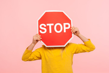 Unrecognizable child in yellow sweatshirt covering face with Stop symbol, anonymous person holding red traffic sign, warning about road safety rules. indoor studio shot isolated on blue background - obrazy, fototapety, plakaty