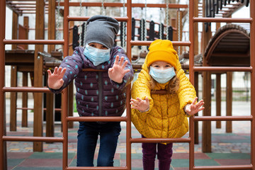 Fototapeta na wymiar two girls in medical masks look at the camera and wave their hands at the playground in coronavirus quarantine period. European pandemic coronavirus concept