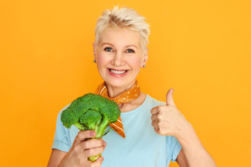 Attractive middle aged woman with short pixie hair holding fresh broccoli and smiling at camera...