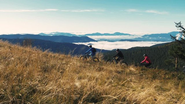 Beautiful aerial view of friends cycling on a narrow mountain path with an amazing view to a foggy forest on a sunny morning day. MTB E-bikes for healthy cycling lifestyle.