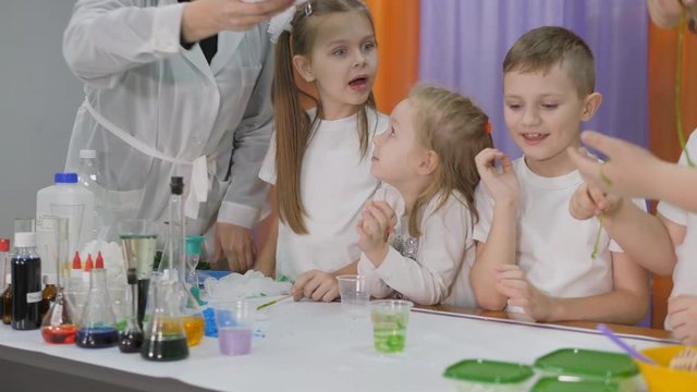 Chemical experiments for children. Woman pours a green substance into a clear glass. Children are surprised. The girl is holding a long green worm. Room is filled with artificial smoke.