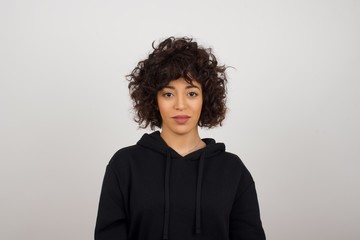 Close up studio shot of beautiful young mixed race woman model with curly dark hair looking at camera with charming cute smile while posing against white blank copy space wall for your content