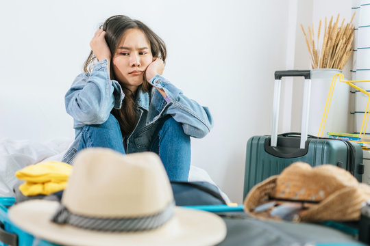 young asian pretty woman hipster traveler sad after flight cancelled with packing clothes into suitcase for holiday vacation on bed at home, delayed flight, summer holiday, backpacker, travel concept