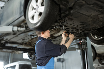 Handsome mechanic in uniform is working in auto service with lifted vehicle. Car repair and maintenance. Oil change