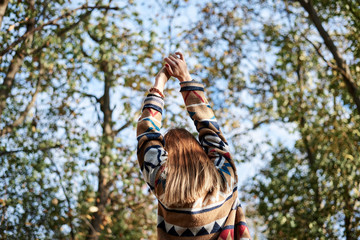 Young blond woman, wearing colorful cardigan, stretching her arms up to the sun in park in autumn. Close-up picture of girl from the back in front of blue sunny sky and green yellow trees in forest.