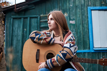 Young blond woman, wearing colorful cardigan, holding acoustic guitar, sitting on a bench in front...