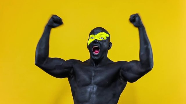 Man sports fan celebrates team victory or goal. Bodybuilder athlete with yellow color on face art and black body paint. Colorful portrait of the guy with bodyart. Slow motion.