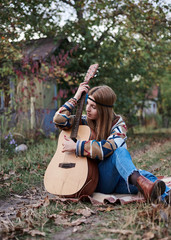Fototapeta na wymiar Young blond woman with bracelets on her hands wearing colorful cardigan and blue jeans, holding embracing acoustic guitar, sitting on ground in countryside. Hippie musician with guitar outside in park