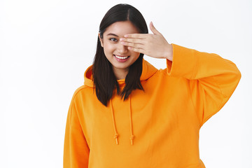 Waist-up portrait beautiful asian modern girl in stylish orange hoodie, cover one eye with palm and smiling, promo of beauty skincare products and korean makeup, standing white background