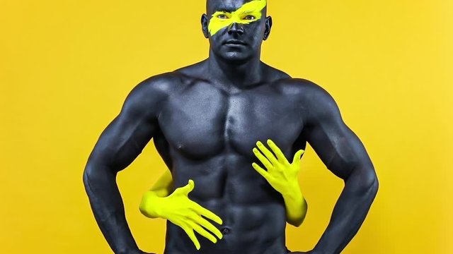 Man with female hands on the body. Bodybuilder athlete with yellow face art and black body paint. Colorful portrait of the guy with bodyart. Slow motion.