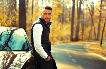 A handsome young Caucasian man wearing style black vest standing near black car on nature.