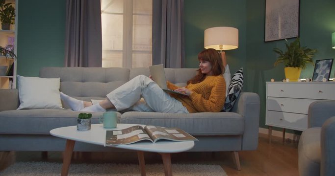 Caucasian female freelancer lying on sofa at home and working on laptop computer. Pretty red-haired woman typing on keyboard and surfing online in cozy living room.