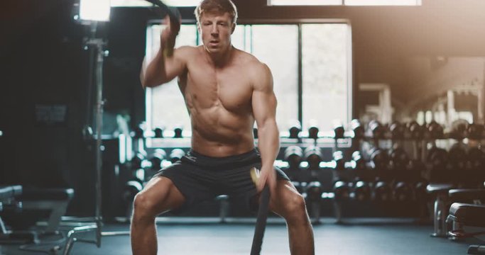 Attractive muscular man working out in the gym using battle ropes with high intensity, determined fitness man achieving his goals