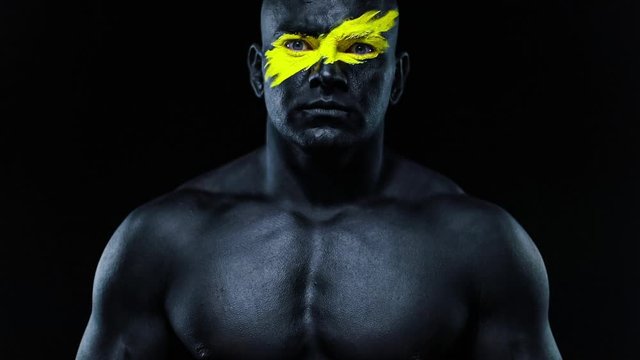 Closeup portrait of man bodybuilder athlete with yellow color on face art and black body paint. Colorful photo of the guy with bodyart. Slow motion.