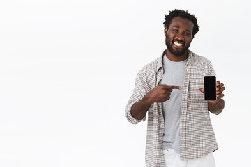 Friendly african-american handsome guy in checked shirt, showing smartphone screen, pointing mobile display and smiling satisfied, sharing cool link or application, recommend download something