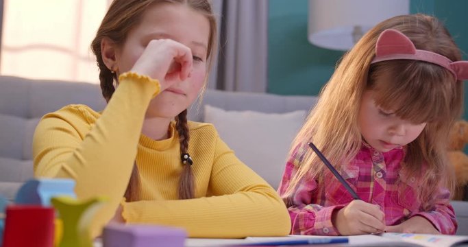 Close up of cheerful small Caucasian sisters sitting at home alone and playing nice together. Cute kids coloring picture with colorful pencils in cozy room. Happy children play and chatting indoor.