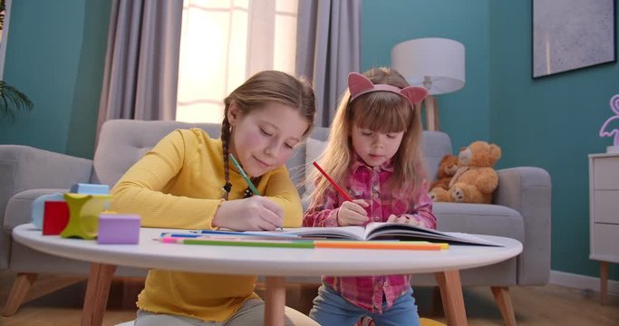 Caucasian little cute kids sitting in cozy nice room at table and coloring pictures with colorful pencils. Cheerful children having fun and play indoors. Girls staying home alone.