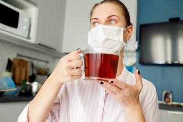 Sick girl in medical mask looking at side with mug of tea in hand standing in kitchen in apartment .