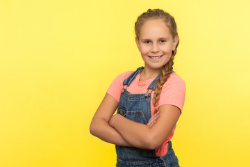 Portrait of cheerful little girl with braid in denim overalls holding hands crossed and smiling at camera, happy child expressing confidence optimism. indoor studio shot isolated on yellow background
